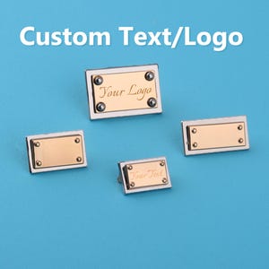 Op de afbeelding: Vier rechthoekige metalen labels met een goudkleurig midden en een zilverkleurige rand. Elk label heeft vier kleine, donkere klinknagels. De labels hebben de tekst "Custom Text/Logo", "Your Logo" en "Your Text". De achtergrond is effen lichtblauw.