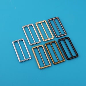 Pode incluir: Uma coleção de deslizadores retangulares de metal em vários acabamentos, incluindo prata, ouro, bronze e preto. Cada deslizador apresenta uma barra central e duas barras laterais, contra um fundo azul brilhante. Estes deslizadores são provavelmente usados para ajustar correias.