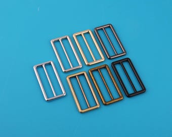 May include: A collection of rectangular metal slides in various finishes, including silver, gold, bronze, and black. Each slide features a central bar and two side bars, set against a bright blue background. These slides are likely used for adjusting straps.