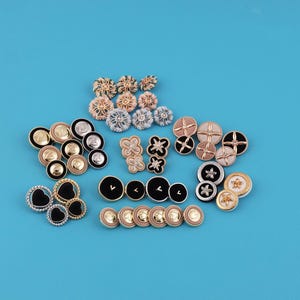 May include: A collection of decorative buttons in various shapes and colors, including gold, black, silver, and cream. The buttons feature floral, heart, and geometric designs, with a variety of textures and finishes. The buttons are arranged on a solid blue background.