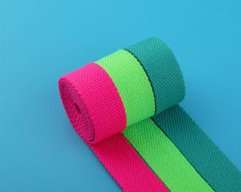 May include: A close-up shot of three rolls of colorful webbing. The rolls are in bright pink, neon green, and teal. The webbing is made of a tightly woven fabric. The background is a solid light blue.