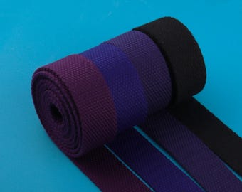 May include: A roll of woven fabric in shades of purple and black. The fabric is tightly woven with a textured appearance. The roll is partially unraveled, revealing the different color stripes. The background is a solid blue.