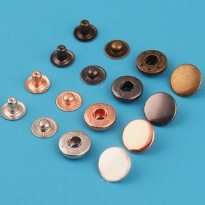 May include: A collection of metal snap fasteners in various finishes including antique bronze, copper, silver, and gunmetal. The set includes circular button caps, socket components, and stud pieces with smooth and textured edges arranged on a surface.