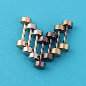 May include: An assortment of barbell-shaped stud earrings in gold, silver, and gunmetal colors. The earrings are arranged on a bright blue surface, showcasing their metallic sheen and simple design. These earrings are suitable for everyday wear.