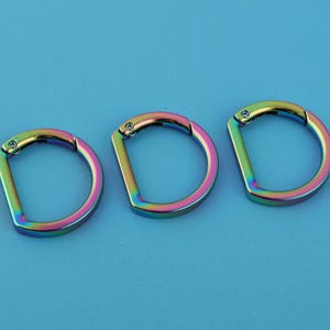 Puede incluir: Tres anillas en D iridiscentes con acabado arcoíris. La ferretería metálica tiene forma de D con una abertura articulada. Las anillas están sobre un fondo azul claro liso. Adecuado para diversos proyectos de artesanía y accesorios.