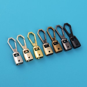 Pode incluir: Uma variedade de sete puxadores de zíper de metal em vários acabamentos metálicos, incluindo prata, ouro e preto. Cada puxador tem uma base retangular com um pequeno orifício e uma argola hexagonal. Dispostos sobre um fundo azul liso.