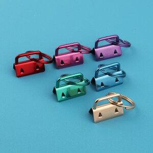 Pode incluir: Uma coleção de ferragens coloridas de metal para chaveiros em vermelho, rosa, roxo, verde, azul e dourado. Cada peça tem uma base retangular com dois orifícios triangulares e uma alça, presa a um anel dividido. A ferragem está em um fundo azul sólido.