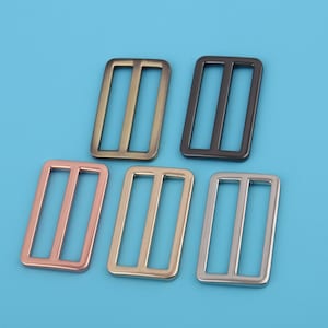 May include: Five metal slider buckles in different finishes: gold, silver, rose gold, black, and antique brass. Each buckle is rectangular with two parallel slots.