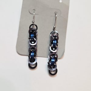 Puede incluir: Un par de pendientes hechos a mano con un diseño de cota de malla. Los pendientes están compuestos por pequeños anillos metálicos entrelazados en plata, negro y azul. Los pendientes cuelgan de ganchos plateados y se exhiben en una tarjeta gris.