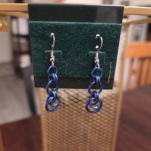 Może przedstawiać: Para niebieskich kolczyków z łańcuszka. Każdy kolczyk ma srebrny haczyk i szereg połączonych ze sobą niebieskich metalowych pierścieni. Kolczyki są prezentowane na ciemnozielonej aksamitnej powierzchni.
