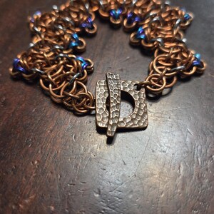 May include: A copper-toned chainmail bracelet with a square clasp. The bracelet features a woven design with small blue beads interspersed throughout. The clasp has a textured surface. The bracelet is displayed on a dark brown surface.