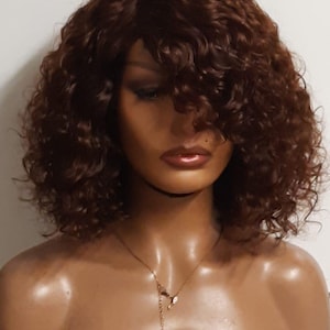 May include: A brown curly wig with a side part. The wig is on a mannequin head with a gold necklace.