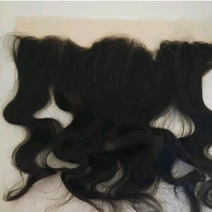 以下が含まれることがあります： ウェーブのかかった黒色のヒューマンヘアーレースフロント。レースフロントは白い表面の上に置かれています。