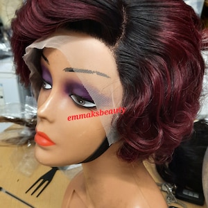 May include: A short, wavy burgundy and black wig with a lace front. The wig is styled in a side part with the burgundy color on the bottom and the black color on the top. The wig is on a mannequin head.