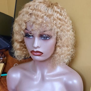 May include: A blonde, curly wig with a short, bob-style cut is displayed on a mannequin head. The wig has a natural-looking hairline and is styled with soft, voluminous curls. The mannequin has a neutral skin tone and is set against a yellow background.