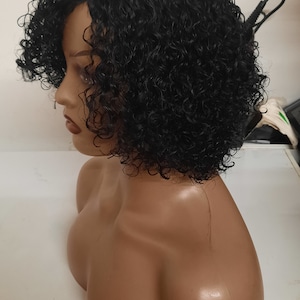 Op de afbeelding: Een zwart krullend pruik op een mannequinhoofd. De pruik is kort en heeft een scheiding aan de zijkant. De mannequin draagt een bruin lichaam.