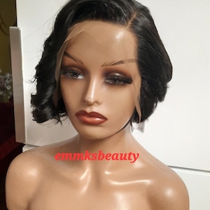 Può includere: Una parrucca nera, corta e a strati, con frangia laterale e riccioli morbidi. La parrucca è su una testa di manichino con un tono di pelle naturale. Il testo "emmsbeauty" è visibile sul petto del manichino.
