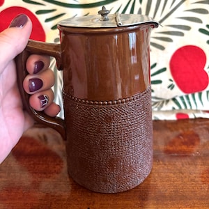May include: A brown ceramic jug with a silver lid. The jug has a textured lower section and a handle. The lid features a small decorative knob. The jug is held in a hand.