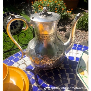 May include: An antique silver teapot with detailed floral and scroll patterns. The teapot features a curved spout, a decorative handle, and a lid with a floral finial. It rests on a blue and white tiled table, with yellow crockery visible.