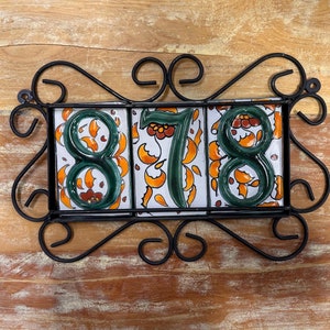 May include: Black metal house number holder with three white ceramic tiles with green and orange floral designs. The numbers 878 are painted in green on the tiles.
