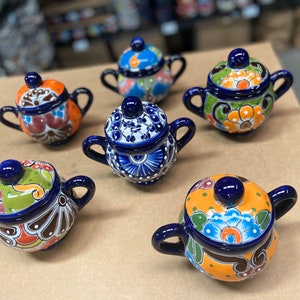 May include: Six small ceramic sugar bowls with lids. Each bowl is hand-painted with colorful floral designs and blue accents. The bowls are perfect for adding a touch of Mexican charm to your kitchen.