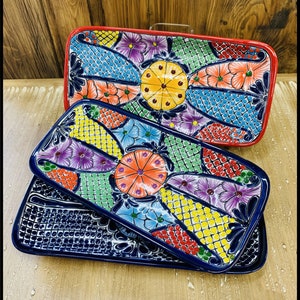 May include: Three colorful ceramic serving trays with a floral and geometric design. The trays are stacked on top of each other. The top tray is red, the middle tray is green, and the bottom tray is blue.
