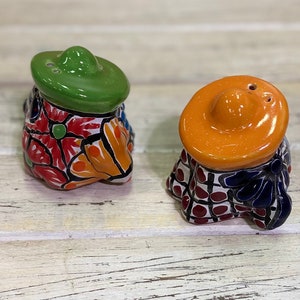 May include: A pair of ceramic salt and pepper shakers. One shaker has a green sombrero, the other has an orange sombrero. Both shakers have a colorful floral design with red, orange, and blue accents. The shakers are on a white wooden surface.