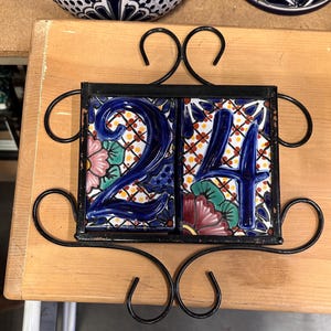 May include: A black metal frame with a decorative scroll design holds two ceramic tiles with the number 24 painted in blue on a colorful floral background.