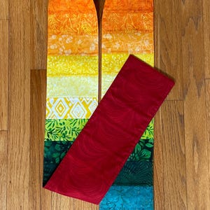May include: A long, rectangular fabric stole featuring a vibrant rainbow gradient of horizontal patchwork segments. The colors transition from deep red and orange to yellow, green, blue, and purple. A solid dark red fabric panel overlays the center.