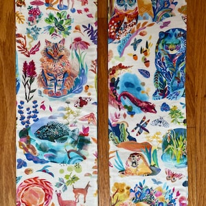 May include: Two long fabric strips featuring a white background with a colourful, repeating pattern of forest animals including deer, a cat, an owl, a bear, and a beaver, interspersed with mushrooms, insects, flowers, and various woodland foliage.
