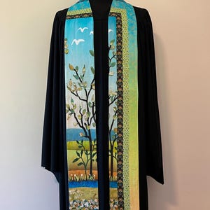 May include: A liturgical stole featuring a landscape scene with trees, white birds, and a blue sky. The design includes a patterned border with floral motifs in shades of green, orange, and black, layered over a solid black robe on a mannequin.