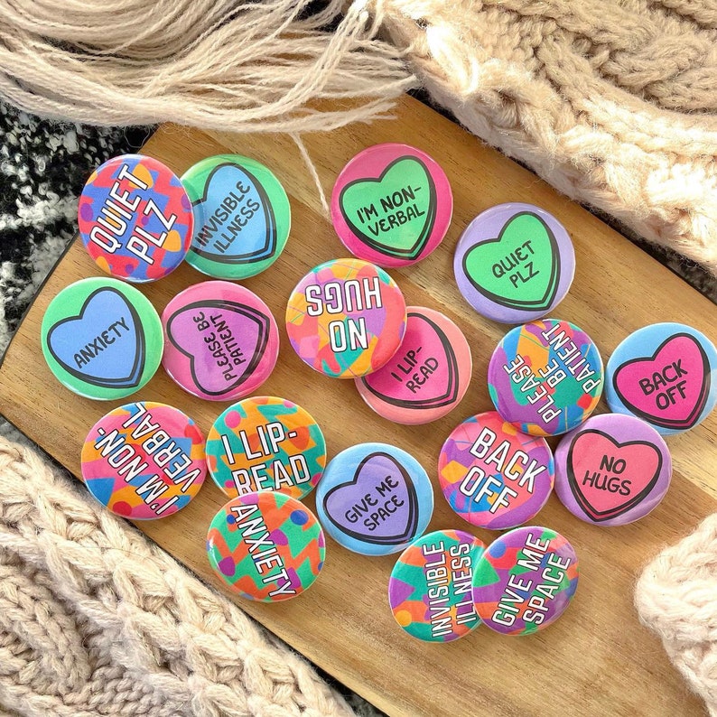 Pu&ograve; includere: Una collezione di 14 bottoni rotondi colorati con vari messaggi stampati su di essi. I messaggi includono "Quiet Plz", "I'm Non-Verbal", "Hugs On", "I Lip-Read", "Anxiety", "Invisible Illness", "Please Be Patient", "Give Me Space", "Back Off", "No Hugs", e altro ancora.