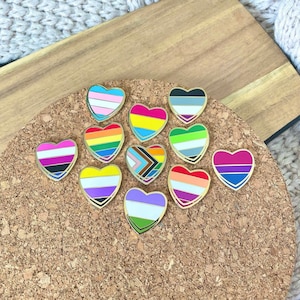 May include: A set of 12 heart-shaped pins featuring various pride flags, including the transgender, lesbian, gay, bisexual, pansexual, and asexual flags. The pins are gold-toned and have a glossy finish.