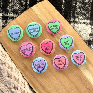 Pu&ograve; includere: Nove bottoni a forma di cuore con messaggi diversi. I messaggi includono "Ansia", "Malattia invisibile", "Sono non verbale", "Per favore, sii paziente", "Leggo sulle labbra", "Dammi spazio", "No abbracci", "Silenzio per favore", e "Vattene".