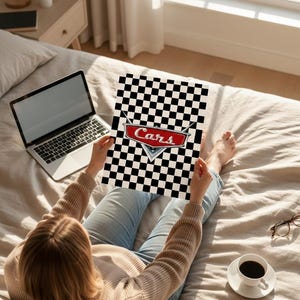May include: A person holding a poster with a black and white checkered pattern and the word "Cars" in red and white. The poster is held over a bed with a laptop, coffee, and glasses nearby.