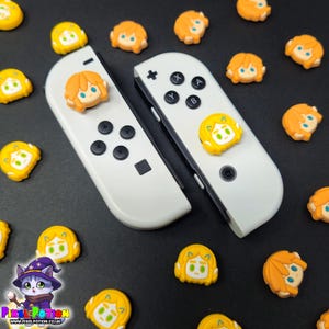 May include: White Nintendo Switch Joy-Con controllers with character-themed thumb grips. The thumb grips are yellow and orange, with a cartoon character design. The controllers are surrounded by additional character thumb grips on a black surface.