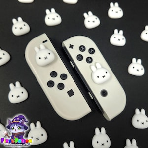 May include: Off-white Nintendo Switch Joy-Con controllers with white bunny face accents and black buttons. Small white bunny face decorations are scattered on a black surface. The controllers have a cute, playful design.
