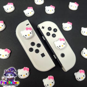 May include: White Nintendo Switch Joy-Con controllers with Hello Kitty face embellishments and scattered Hello Kitty face charms with pink bows on a black surface. The controllers have black buttons and a black rail.