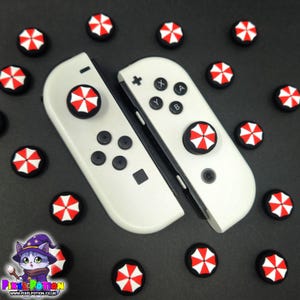 May include: White game controllers with black buttons and red and white umbrella-themed thumb grips. Additional thumb grips with the same design are scattered around. The image is on a black background.
