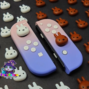 May include: Two Nintendo Switch Joy-Cons with a pink and purple gradient design, featuring bunny-shaped buttons. Additional bunny-shaped buttons in white and brown are scattered around the controllers. The controllers have white buttons and a black frame.