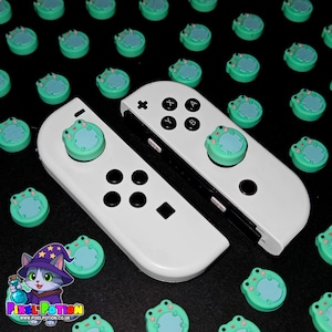 May include: White game controller with black buttons and frog-shaped thumb grips in light green and blue. Additional frog thumb grips are scattered around the controllers. The Pixel Potion logo is in the lower left corner.