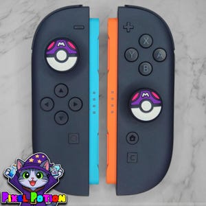 May include: Two dark grey game controllers with a light blue side and an orange side. Each controller features a circular joystick cover designed like a purple, pink, and white ball with an M symbol. The controllers include standard buttons.