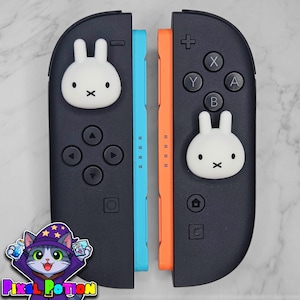 May include: Two black game controller attachments featuring white rabbit-shaped joystick covers. The left controller has a light blue side rail, while the right controller has an orange side rail. Each controller includes standard directional buttons, action buttons, and a home button.