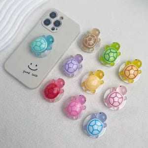 May include: A selection of colourful turtle-shaped phone grips. The grips are in various colours, including blue, green, pink, purple, and yellow. One is attached to a white phone case with the text "good luck" and a smiley face.
