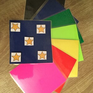 Könnte beinhalten: Ein Stapel aus acht bunten Blättern Papier mit fünf gelben Sternen auf dem oberen Blatt. Die Farben der Blätter sind Schwarz, Blau, Grün, Limettengrün, Gelb, Rot und Pink.