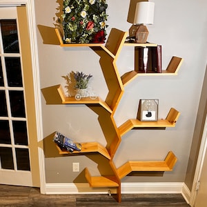 Op de afbeelding: Een houten boekenkast in de vorm van een boom met meerdere planken. De planken zijn in een uniek, organisch patroon gerangschikt, dat lijkt op takken. De boekenkast is aan een muur bevestigd en heeft een kleine potplant, een ingelijst beeld en een lamp op de bovenste plank.