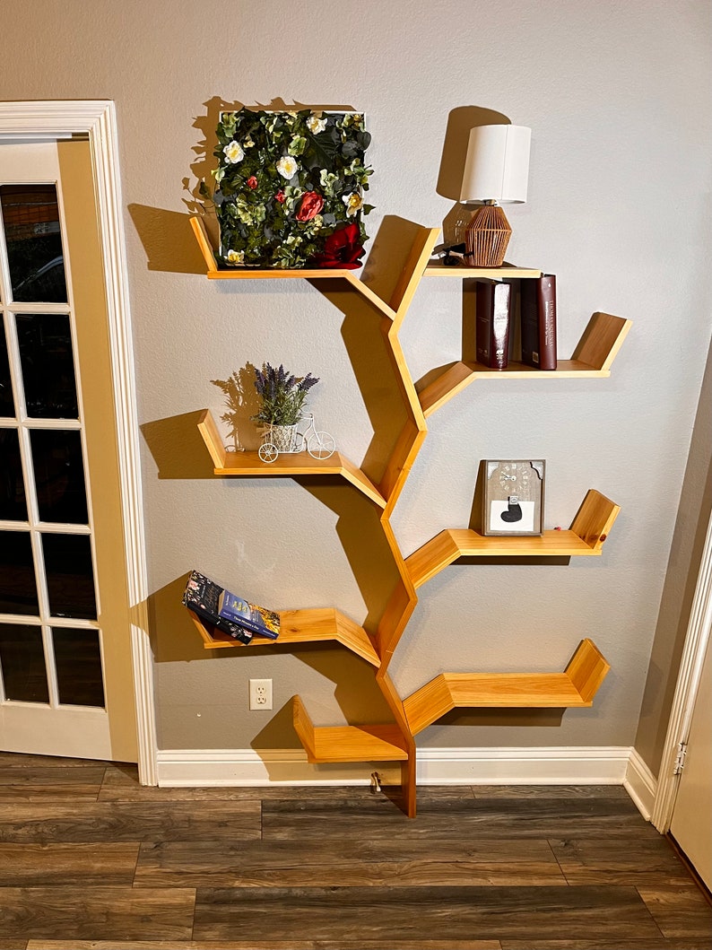 Op de afbeelding: Een houten boekenkast in de vorm van een boom met meerdere planken. De planken zijn in een uniek, organisch patroon gerangschikt, dat lijkt op takken. De boekenkast is aan een muur bevestigd en heeft een kleine potplant, een ingelijst beeld en een lamp op de bovenste plank.