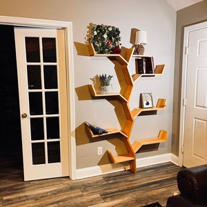 Op de afbeelding: Een houten boekenkast in de vorm van een boom met meerdere planken. De planken zijn gevuld met boeken, planten en decoratieve items. De boekenkast is gemonteerd aan een lichtbeige muur.