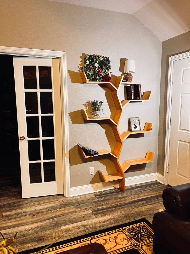 Op de afbeelding: Een houten boekenkast in de vorm van een boom met meerdere planken. De planken zijn gevuld met boeken, planten en decoratieve items. De boekenkast is gemonteerd aan een lichtbeige muur.