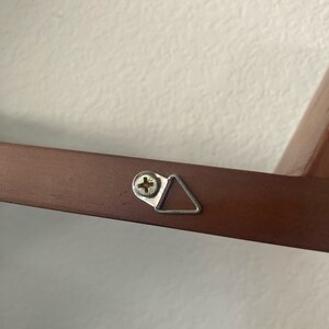Op de afbeelding: Een close-up van een bruin houten frame met een zilveren driehoekige ophanghaak bevestigd met een schroef. De haak is ontworpen om een ​​foto of ander object aan een muur te hangen.